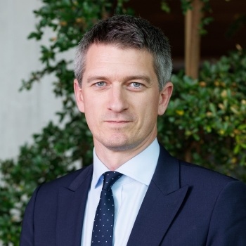 Tom has short greyish hair. He is wearing a navy suit and tie