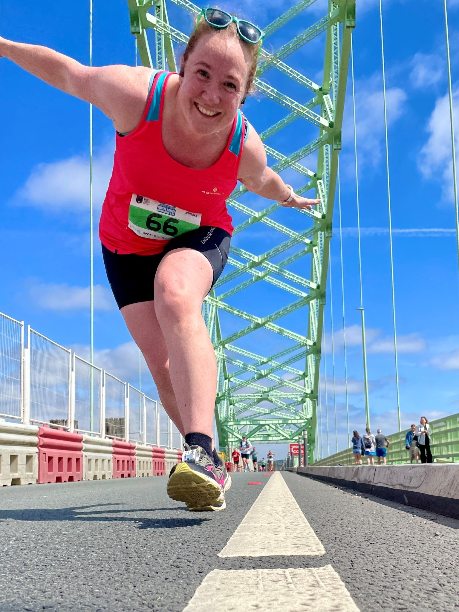 Alison is pictured during a 10k race on Jubilee Bridge in London. She is bending down towards the camera with her arms spread out while running and is smiling.