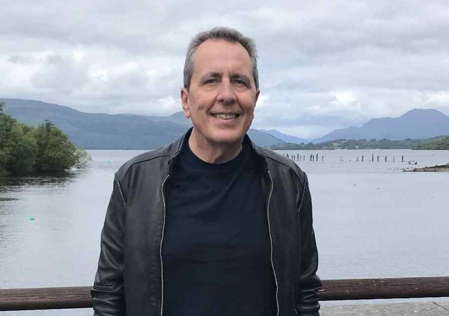 Bill is standing in front of a lake with mountains behind. He has short hair