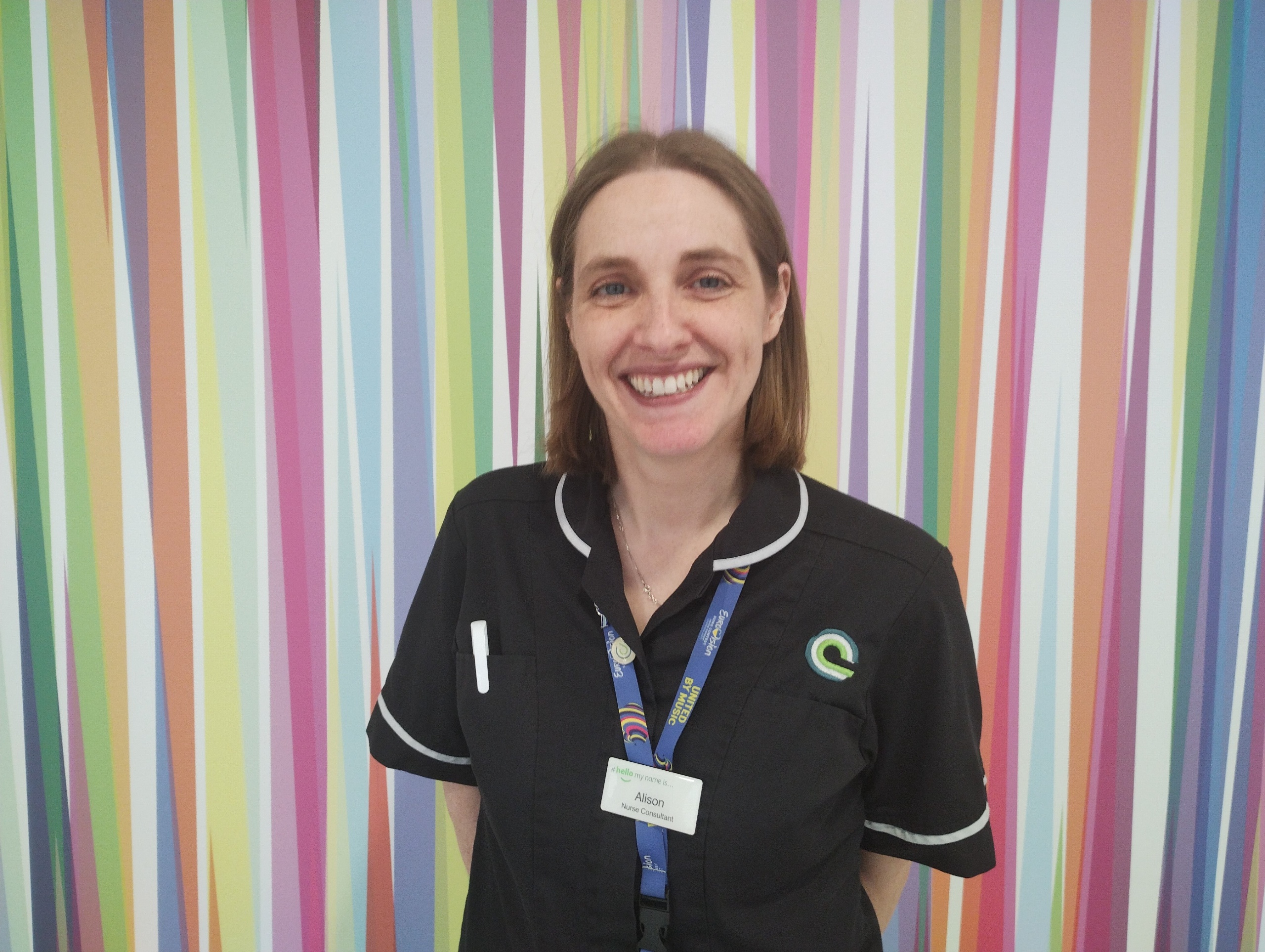 Alison wears a navy nurses tunic and has shoulder length brown hair. She is standing in front of brightly striped artwork on the walls of the hospital outpatients department