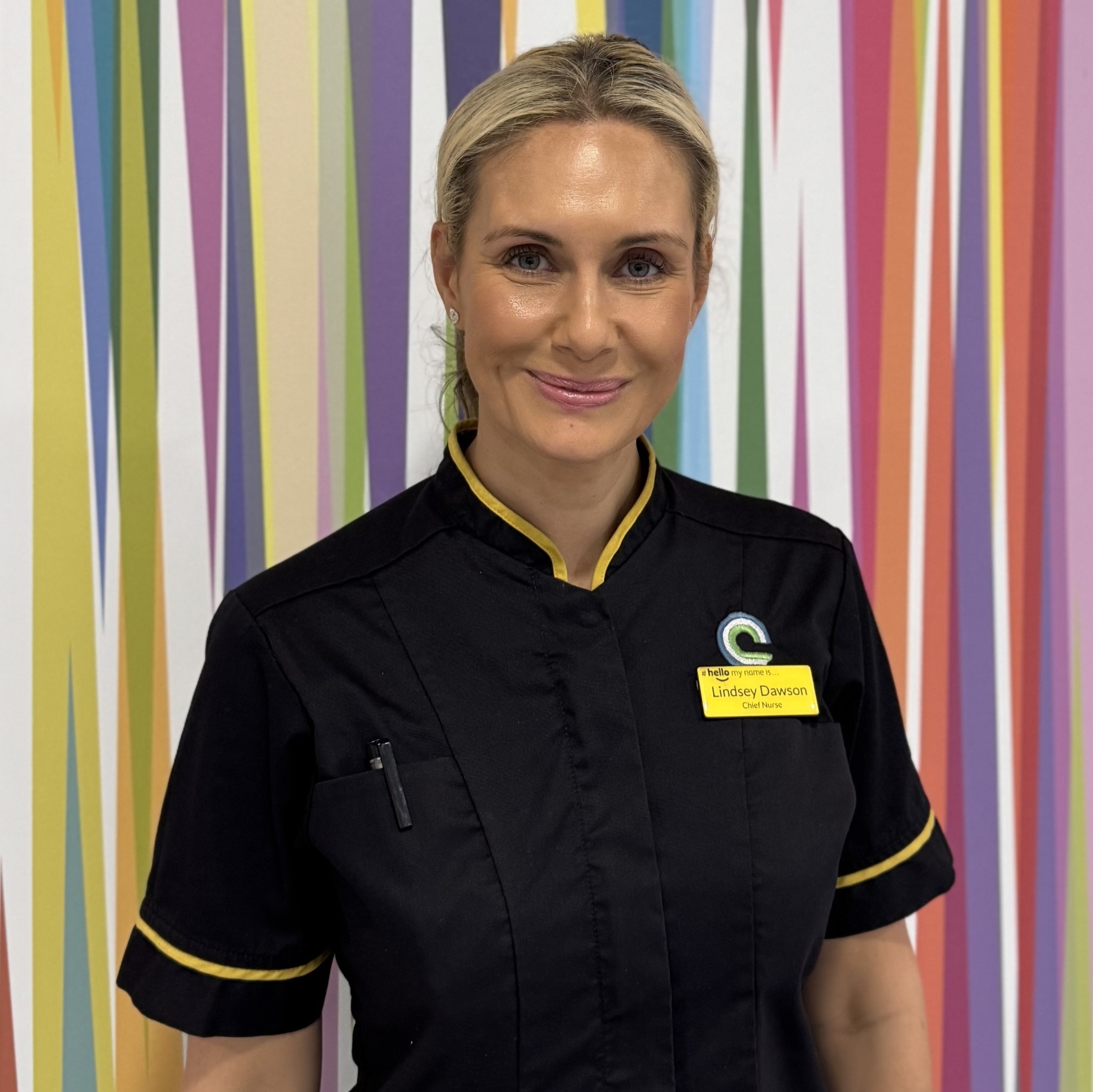 Lindsey has blonde hair tied back and is wearing a black nurses dress with yellow piping