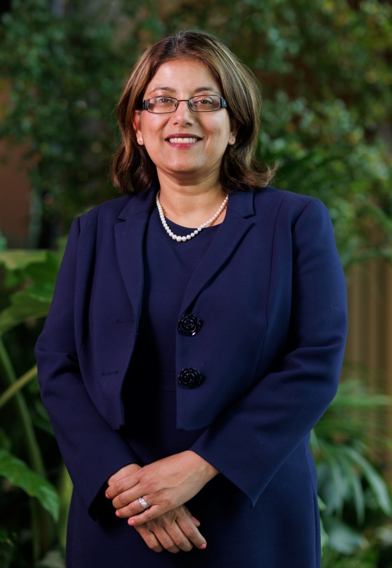 Sheena has brown hair in a shoulder-length bob and is wearing a dark navy dress and jacket and glasses. She is standing in a lobby with lots of plants and greenery behind her