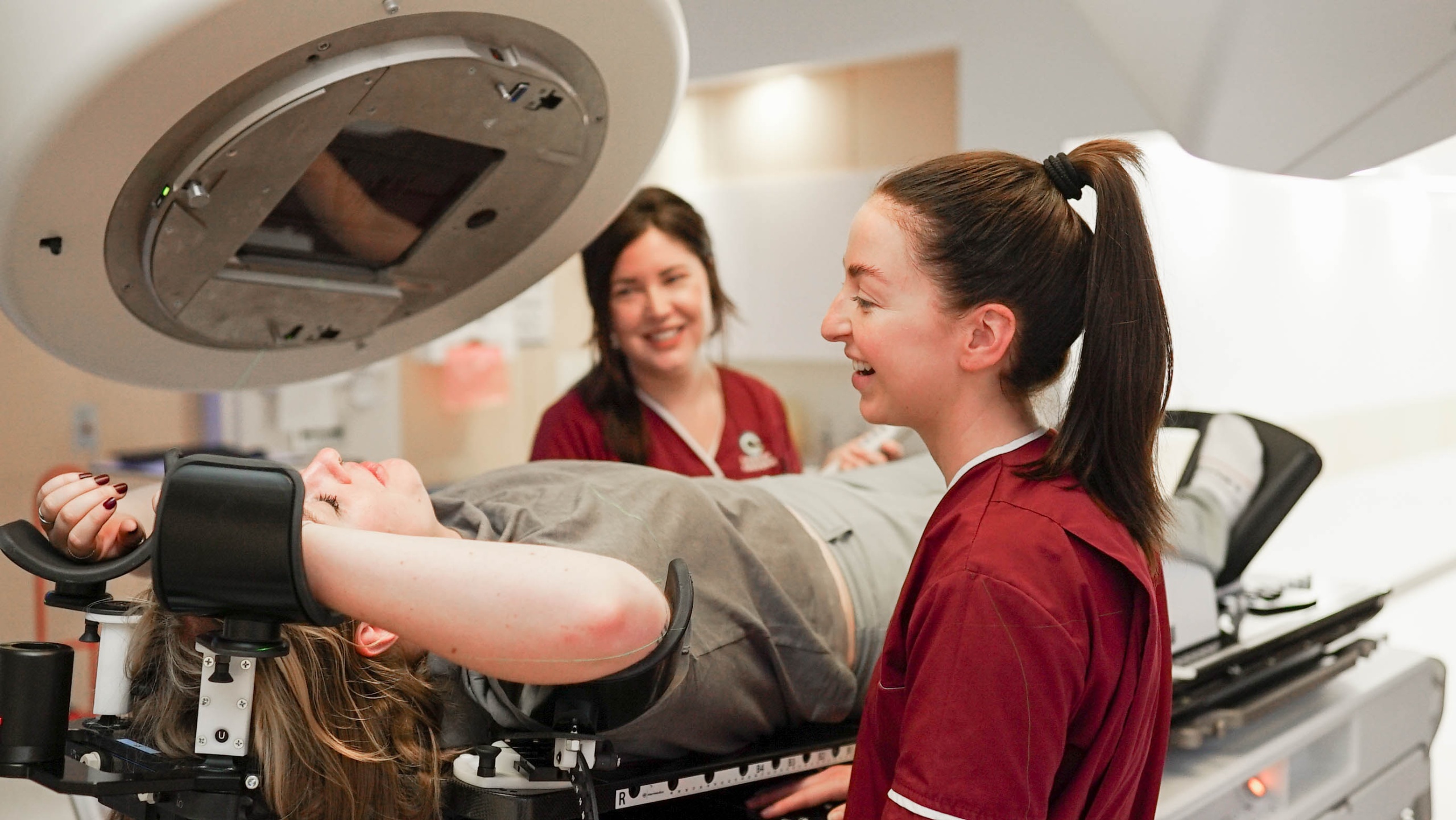 Two therapeutic radiographers in burgundy scrubs with a patient lying on a radiotherapy treatment machine, known as a linac