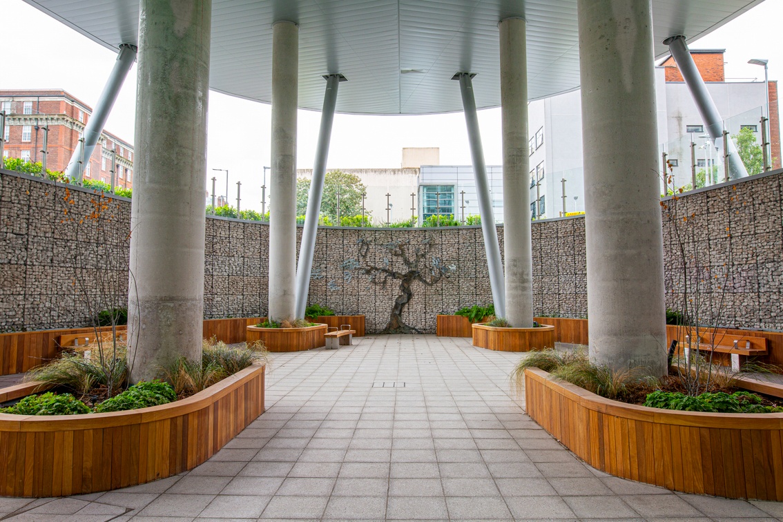 An outdoor area with paving and plants. It is covered by a canopy supported by pillars