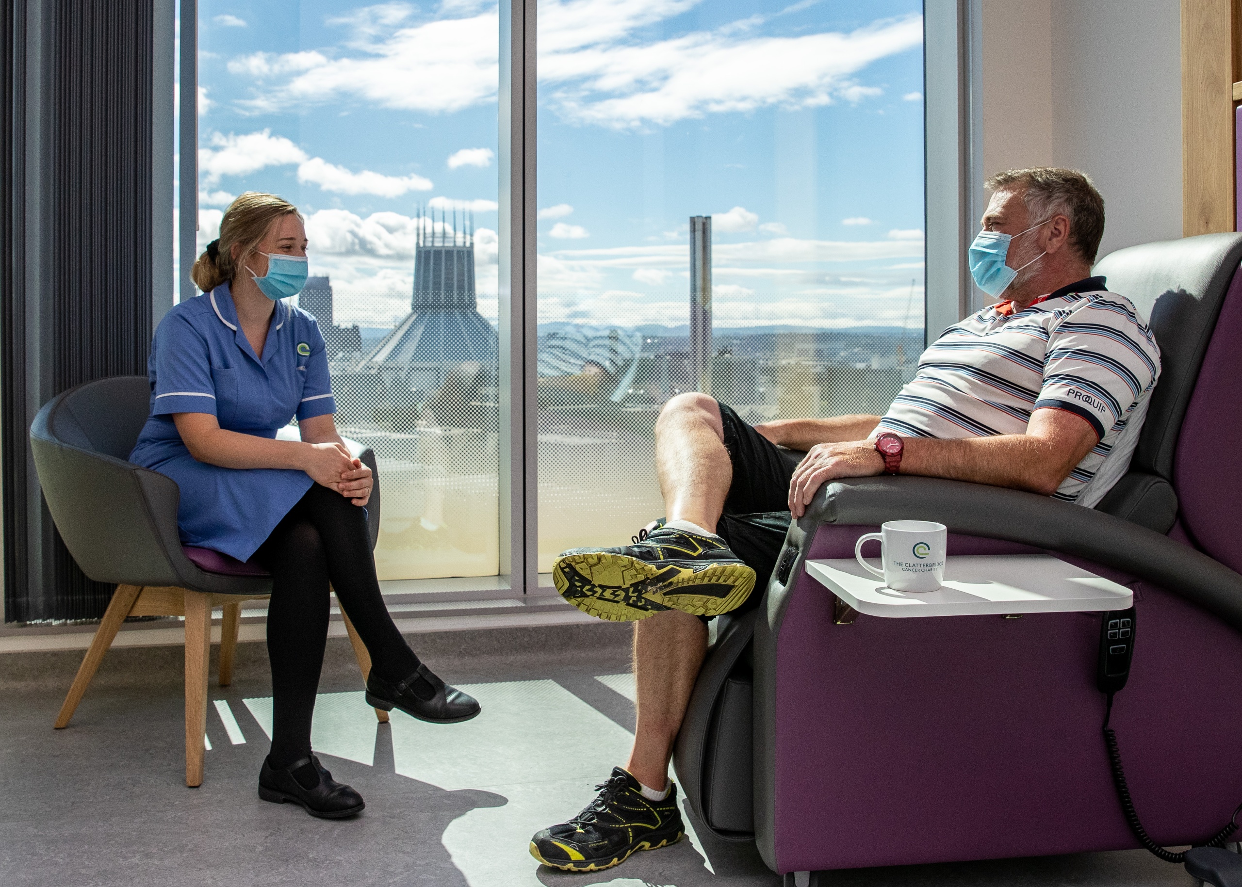 Male patient relaxing in chemothgerapy treatment chair, chatting to a female nurse sitting next to him. Liverpool Metropolitan Cathedral is in the background, through the window