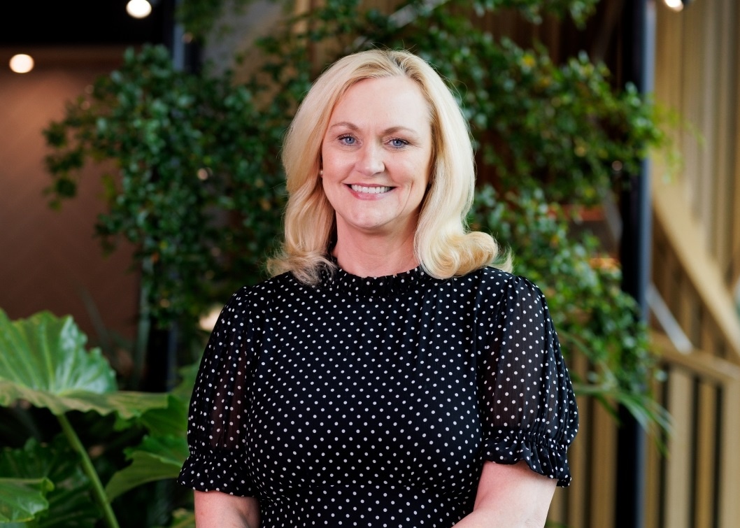 Joan has blonde hair and is standing in a reception area with green plants around. She is smiling